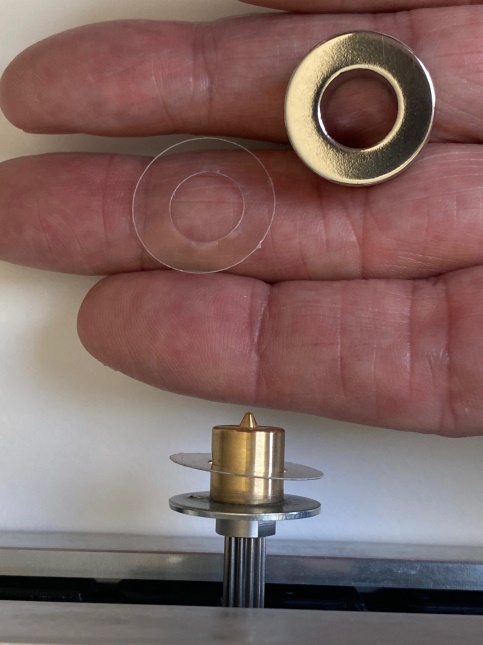 Photo of a hand holding the ring-shaped magnet and a thin plastic shim, ready to be placed on the motor shaft that has the metal washer and drawing tip installed.